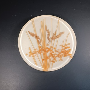 May include: A round, off-white resin coaster with a wheat design. The coaster features dried wheat stalks, long strands, and small orange ring-shaped pieces. The design is set against a white background.