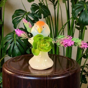 May include: A cream-colored ceramic bird vase with an orange head and yellow beak, holding green and pink floral arrangements. The vase is on a dark brown wooden table, surrounded by green plants.