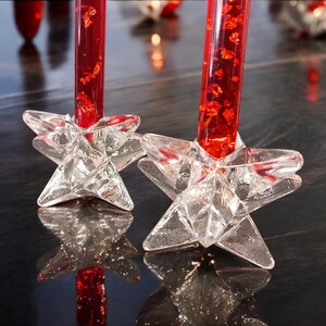 May include: Two clear glass candle holders shaped like five-pointed stars, each supporting a tall, cylindrical red candle filled with suspended red glitter particles. The star-shaped bases feature faceted surfaces that reflect light.