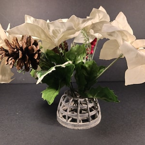 May include: A festive floral arrangement featuring large, off-white poinsettia blooms, green leaves, and a brown pinecone. The flowers are arranged in a gray, cage-like vase, creating a seasonal decorative piece.
