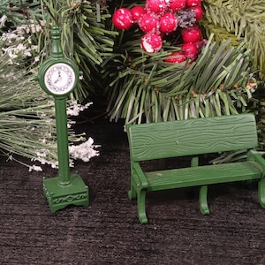 May include: A miniature green park bench with a wood grain texture and a matching green street clock on a pedestal base. The clock features a round white face with black numerals and hands, standing upright next to the bench on a flat surface.