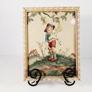 Op de afbeelding: Een ingelijste prent met een kind dat een lam vasthoudt, omringd door andere lammeren en kleine huisjes. Het kunstwerk heeft een vintage esthetiek, met een cr&egrave;mekleurige lijst en een zwarte metalen standaard.