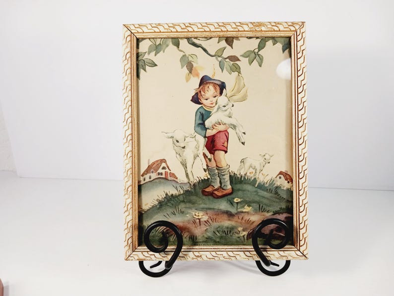 Op de afbeelding: Een ingelijste prent met een kind dat een lam vasthoudt, omringd door andere lammeren en kleine huisjes. Het kunstwerk heeft een vintage esthetiek, met een cr&egrave;mekleurige lijst en een zwarte metalen standaard.