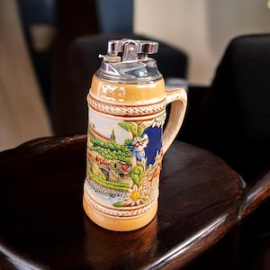 May include: A decorative ceramic lighter in the shape of a beer stein. The stein features a detailed landscape scene with a bridge, buildings, and a river. The lighter has a silver-colored metal top and a handle.