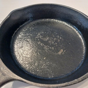 May include: A round black cast iron skillet with a textured interior surface and a pour spout on the side. The cooking surface shows signs of wear with a slightly uneven finish, and the handle extends from the side of the circular pan body.