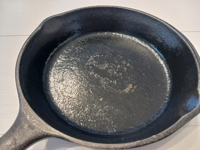 May include: A round black cast iron skillet with a textured interior surface and a pour spout on the side. The cooking surface shows signs of wear with a slightly uneven finish, and the handle extends from the side of the circular pan body.