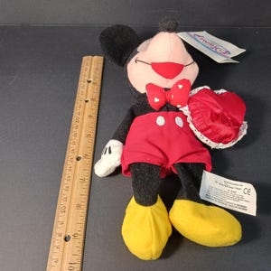 Op de afbeelding: Mickey Mouse pluche speelgoed met een rode strik, rode korte broek en gele schoenen. Het pluche speelgoed houdt een rood hartvormig item met witte kanten rand vast. Een houten liniaal ligt naast het speelgoed.