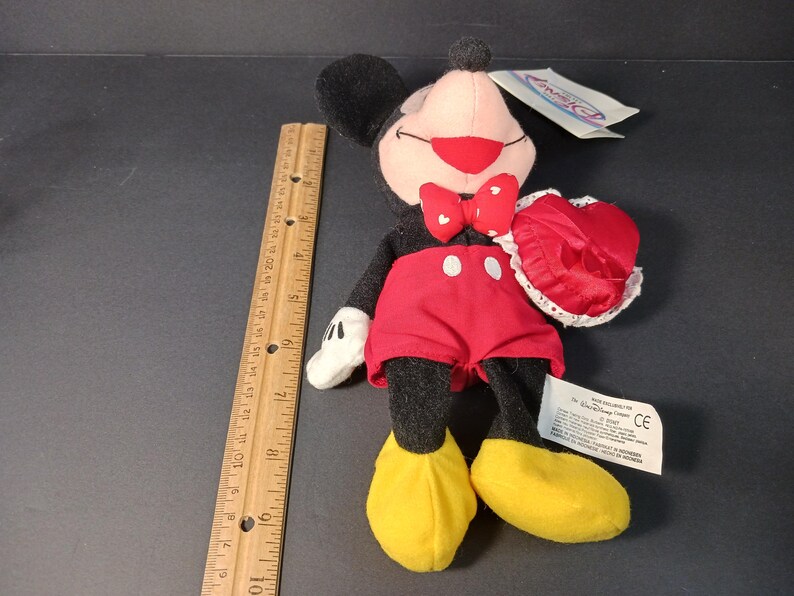 Op de afbeelding: Mickey Mouse pluche speelgoed met een rode strik, rode korte broek en gele schoenen. Het pluche speelgoed houdt een rood hartvormig item met witte kanten rand vast. Een houten liniaal ligt naast het speelgoed.