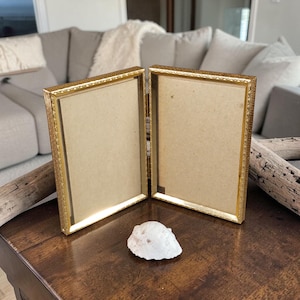May include: A gold-colored, double-hinged picture frame with a decorative border. The frame is open, revealing a tan backing. A white, irregularly shaped stone sits in front of the frame on a dark wooden surface.