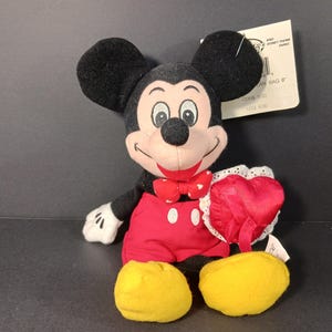 Op de afbeelding: Een Mickey Mouse pluche speelgoed met zwarte oren, een rode vlinderdas en een rode broek. Het pluche houdt een rood hartvormig kussen met witte kanten rand vast. Gele schoenen en witte handschoenen.
