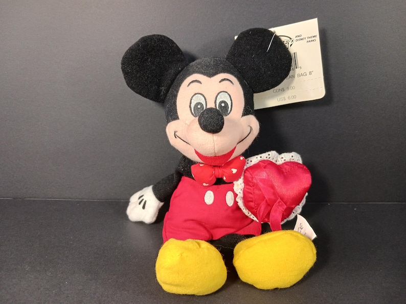 Op de afbeelding: Een Mickey Mouse pluche speelgoed met zwarte oren, een rode vlinderdas en een rode broek. Het pluche houdt een rood hartvormig kussen met witte kanten rand vast. Gele schoenen en witte handschoenen.