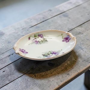 May include: Oval-shaped porcelain dish with a floral design. The dish features a cream-colored base with purple flowers and green leaves. The rim has a gold trim and a light pink border. The dish is placed on a weathered wooden surface.