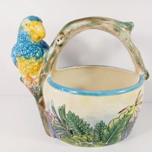 May include: A ceramic basket with a parrot perched on the side. The parrot is blue, yellow, and orange. The basket has a handle and is decorated with a jungle scene. The rim of the basket is trimmed with blue.