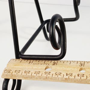 Op de afbeelding: Zwarte metalen decoratieve standaard met een uniek, gebogen ontwerp. Een houten liniaal meet ongeveer 7,6 cm. De poten van de standaard zijn schuin en de bovenkant heeft een decoratieve krul.