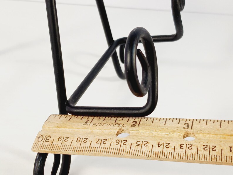 Op de afbeelding: Zwarte metalen decoratieve standaard met een uniek, gebogen ontwerp. Een houten liniaal meet ongeveer 7,6 cm. De poten van de standaard zijn schuin en de bovenkant heeft een decoratieve krul.