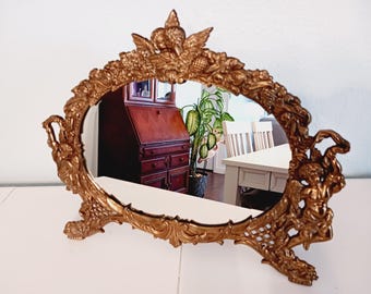 May include: Ornate gold-colored oval vanity mirror. The frame features detailed relief carvings of cherubs, flowers, and foliage. The mirror is supported by two feet and has handles on each side. The mirror reflects a room with furniture and a plant.