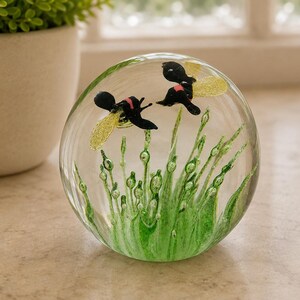 May include: A clear glass paperweight featuring two black bees with shimmering gold wings suspended above a cluster of green glass blades of grass. The bees have small pink accents on their bodies, and the entire scene is encased in a smooth sphere.