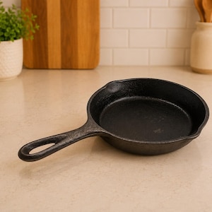 May include: A black cast iron skillet with a round cooking surface, a short handle featuring a circular hole for hanging, and small pouring spouts on the sides. The pan has a textured, matte finish and a deep, circular basin for cooking.