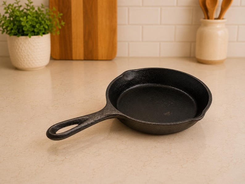 May include: A black cast iron skillet with a round cooking surface, a short handle featuring a circular hole for hanging, and small pouring spouts on the sides. The pan has a textured, matte finish and a deep, circular basin for cooking.