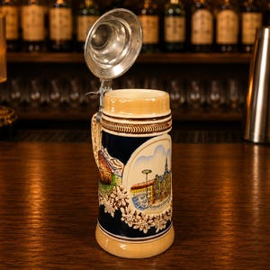 May include: A ceramic beer stein with a hinged metal lid and a handle. The body is dark blue with a cream-colored rim and base, featuring a colorful relief illustration of a cityscape labeled Amsterdam, along with floral accents and a small crest.