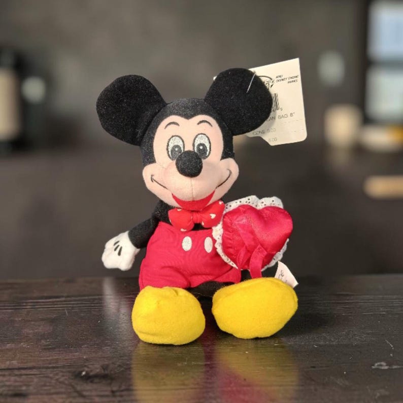 Pode incluir: Um peluche do Mickey Mouse com orelhas pretas, la&ccedil;o vermelho com cora&ccedil;&otilde;es brancos, cal&ccedil;&otilde;es vermelhos e sapatos amarelos. A personagem segura uma almofada vermelha em forma de cora&ccedil;&atilde;o com renda branca. Etiqueta de 20 cm na orelha.