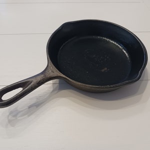 May include: A black cast iron skillet with a round cooking surface, a textured exterior, and a long handle featuring a hole at the end for hanging. The pan includes two small pouring spouts on opposite sides of the rim for liquid drainage.