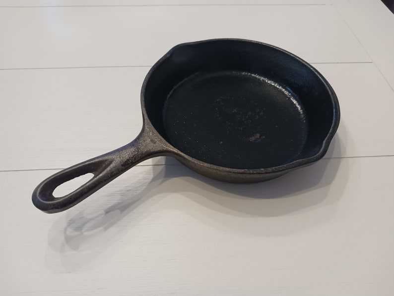 May include: A black cast iron skillet with a round cooking surface, a textured exterior, and a long handle featuring a hole at the end for hanging. The pan includes two small pouring spouts on opposite sides of the rim for liquid drainage.