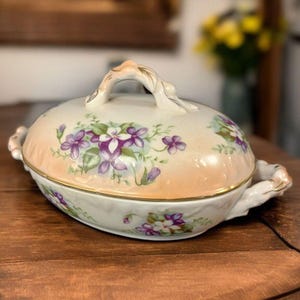 May include: Oval, lidded serving dish with a handle. The dish is cream-colored with a peach-colored band around the middle. It is decorated with purple and white floral designs and gold trim. The dish is likely vintage or antique.