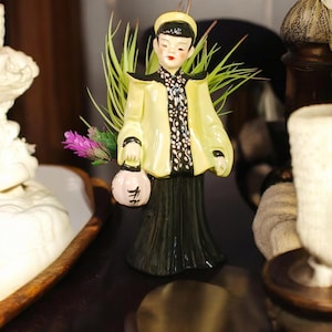 May include: A ceramic figurine of a person in traditional Asian attire. The figure wears a yellow jacket with black trim over a black and white patterned top and a long black skirt. The figure holds a pink lantern and is adorned with greenery and purple flowers.