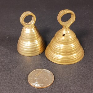 Puede incluir: Dos peque&ntilde;as campanas antiguas de lat&oacute;n con un anillo en la parte superior para colgar. Las campanas tienen forma c&oacute;nica y estriada. Una moneda de veinticinco centavos estadounidense est&aacute; junto a las campanas para comparar el tama&ntilde;o. Las campanas est&aacute;n sobre una superficie oscura.