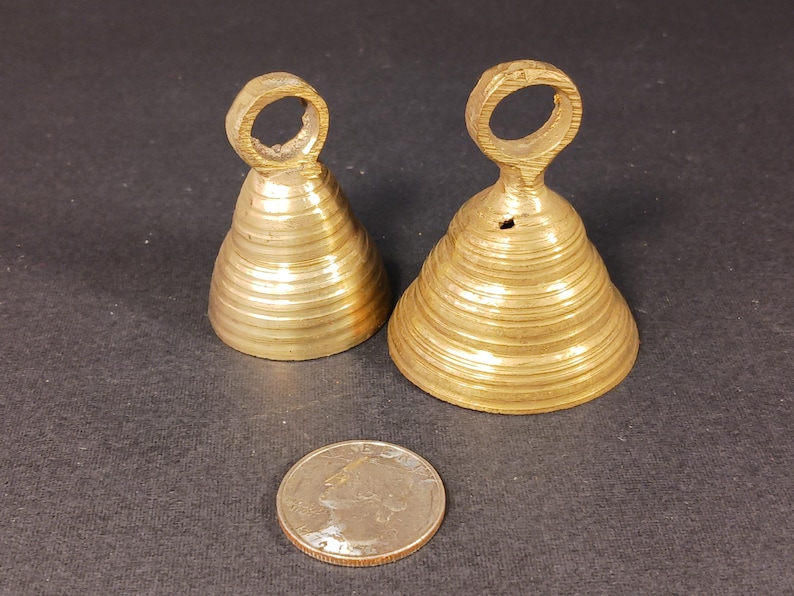 Puede incluir: Dos peque&ntilde;as campanas antiguas de lat&oacute;n con un anillo en la parte superior para colgar. Las campanas tienen forma c&oacute;nica y estriada. Una moneda de veinticinco centavos estadounidense est&aacute; junto a las campanas para comparar el tama&ntilde;o. Las campanas est&aacute;n sobre una superficie oscura.
