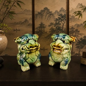May include: Two ceramic Foo Dog statues. The statues are green, blue, and yellow with detailed features. They are positioned on a dark surface, with a traditional Asian-style painting in the background. Bamboo and a potted plant are also visible.