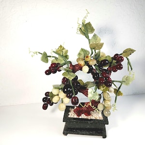May include: A decorative artificial grape arrangement featuring clusters of dark red, light green, and off-white grapes. The grapes are attached to green stems with green leaves. The arrangement sits in a small, dark, rectangular planter.