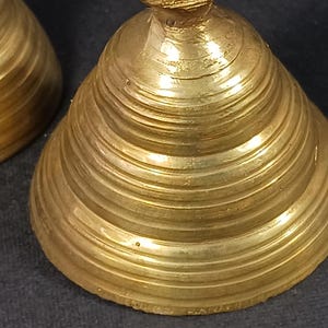 Puede incluir: Primer plano de dos campanas antiguas de lat&oacute;n. Las campanas tienen forma c&oacute;nica con estr&iacute;as horizontales. El lat&oacute;n tiene un tono dorado c&aacute;lido y la superficie muestra signos de edad. Las campanas probablemente se utilizan para decoraci&oacute;n o como instrumentos musicales.