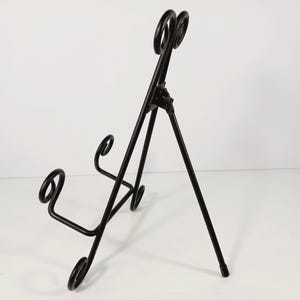 Op de afbeelding: Zwarte metalen displaystandaard met een driepootontwerp. De standaard heeft decoratieve krullen en een bovenkant met twee cirkelvormige accenten. Ideaal voor het presenteren van borden of kunstwerken.