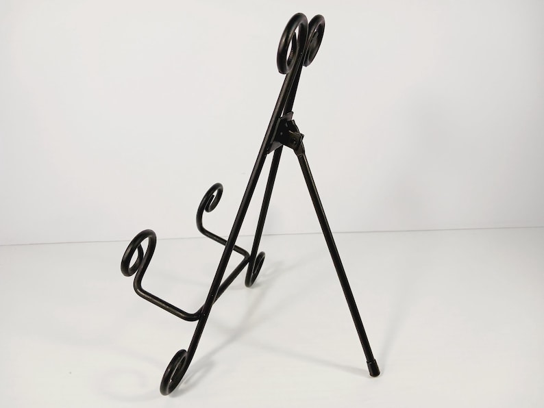Op de afbeelding: Zwarte metalen displaystandaard met een driepootontwerp. De standaard heeft decoratieve krullen en een bovenkant met twee cirkelvormige accenten. Ideaal voor het presenteren van borden of kunstwerken.