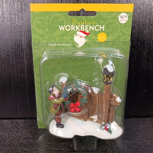 May include: A Santa's Workbench resin accessory in original packaging. The figurine depicts a person in a green coat hanging a green wreath with a red bow on a brown wooden fence next to a black street lamp, all set on a snowy white base.