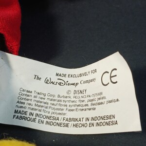 Op de afbeelding: Een close-up van een wit label met tekst, waaronder "MADE EXCLUSIVELY FOR The Walt Disney Company" en "&copy; DISNEY". Het label bevat ook tekst in het Frans, Duits en Spaans. Een CE-markering is zichtbaar.