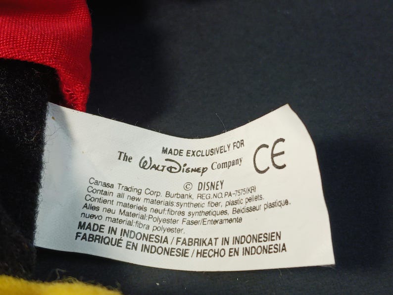 Op de afbeelding: Een close-up van een wit label met tekst, waaronder "MADE EXCLUSIVELY FOR The Walt Disney Company" en "&copy; DISNEY". Het label bevat ook tekst in het Frans, Duits en Spaans. Een CE-markering is zichtbaar.