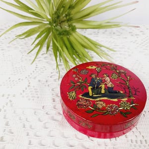 May include: A round, red lacquered box with a detailed illustration on the lid. The illustration depicts two figures in a landscape with floral and tree motifs. The box is on a white lace surface.