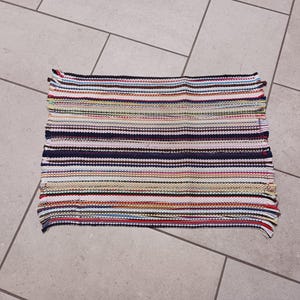 May include: Rectangular, handwoven rug with horizontal stripes in various colors including red, blue, green, and white. The rug has a fringed edge and is likely made of fabric scraps. The rug is placed on a tiled floor.