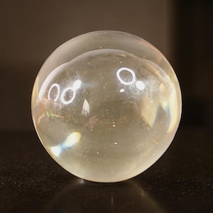 May include: A clear, spherical glass ball with a slightly yellow tint. The ball reflects the surrounding light, creating bright, circular highlights. The surface appears smooth and polished, with subtle imperfections visible. The overall effect is one of clarity and transparency.