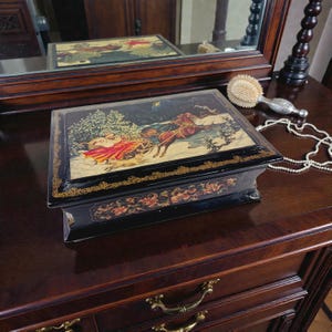 May include: A black decorative box with a winter scene featuring a horse-drawn sleigh. The box has a gold trim and floral accents. The box is sitting on a dark wooden surface.