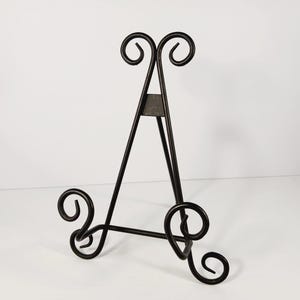 Op de afbeelding: Een zwarte metalen ezel met een decoratief krulontwerp. De ezel heeft een driepotige basis en een driehoekig frame, met krulaccenten aan de boven- en onderkant. De ezel is ontworpen om een fotolijst of kunstwerk vast te houden.