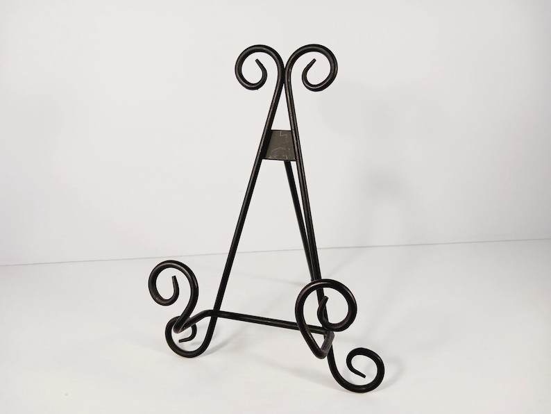 Op de afbeelding: Een zwarte metalen ezel met een decoratief krulontwerp. De ezel heeft een driepotige basis en een driehoekig frame, met krulaccenten aan de boven- en onderkant. De ezel is ontworpen om een fotolijst of kunstwerk vast te houden.