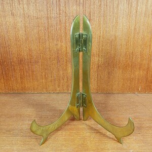 May include: A polished brass display stand featuring two curved legs connected by two central hinges. The stand has a smooth, metallic gold-toned finish and is designed to hold items upright with its two upward-curving support arms at the base.