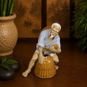 May include: A ceramic figurine of an elderly person with a white beard and hair, wearing a light blue shirt and white pants. The figure is crouching, holding a brown fish in one hand and resting the other hand on a brown, bell-shaped woven basket.