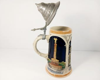 May include: A ceramic beer stein with a hinged metal lid. The stein features a detailed illustration of a monument with the text "Siegessäule" on the front. The handle is cream-colored, and the lid is silver.