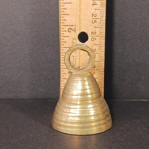 Puede incluir: Una peque&ntilde;a campana antigua de lat&oacute;n con un asa de anillo. La campana tiene forma c&oacute;nica con estr&iacute;as horizontales. Una regla de madera est&aacute; colocada detr&aacute;s de la campana, mostrando medidas en cent&iacute;metros.