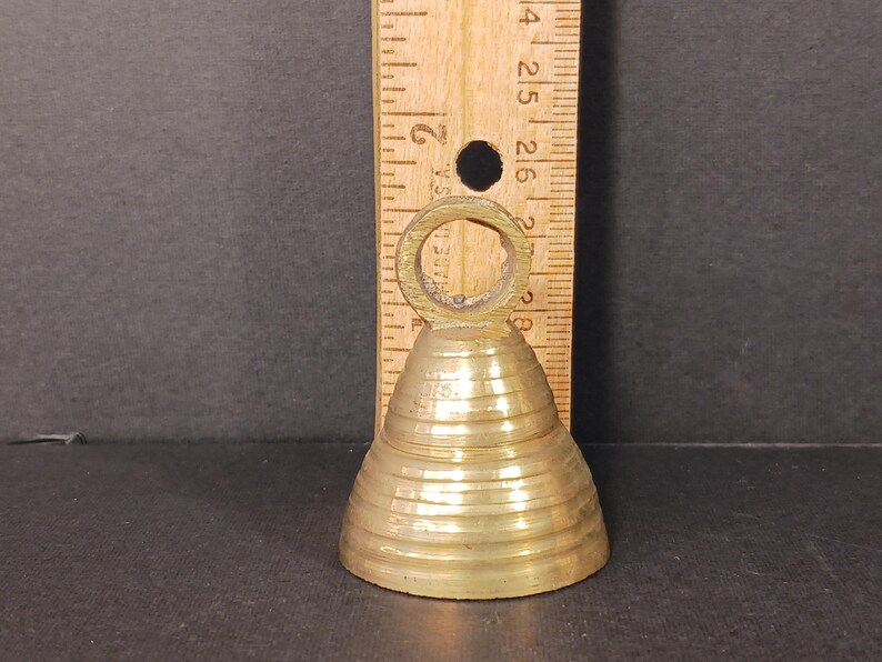 Puede incluir: Una peque&ntilde;a campana antigua de lat&oacute;n con un asa de anillo. La campana tiene forma c&oacute;nica con estr&iacute;as horizontales. Una regla de madera est&aacute; colocada detr&aacute;s de la campana, mostrando medidas en cent&iacute;metros.